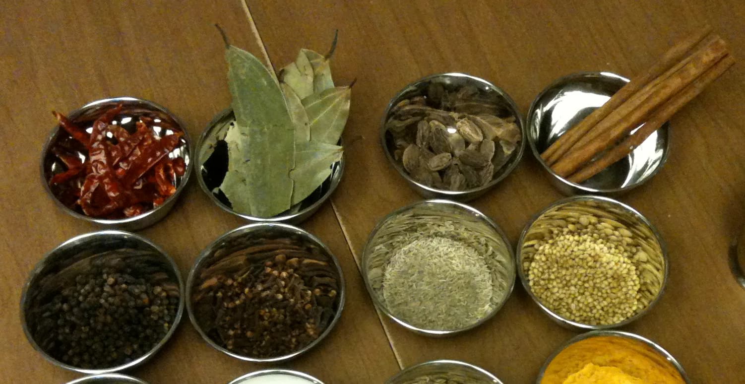 Bowls of whole spices: dried red chillies, bay leaves, black cardamom pods and cinnamon sticks, with black peppercorns, cloves, cumin and coriander seeds below