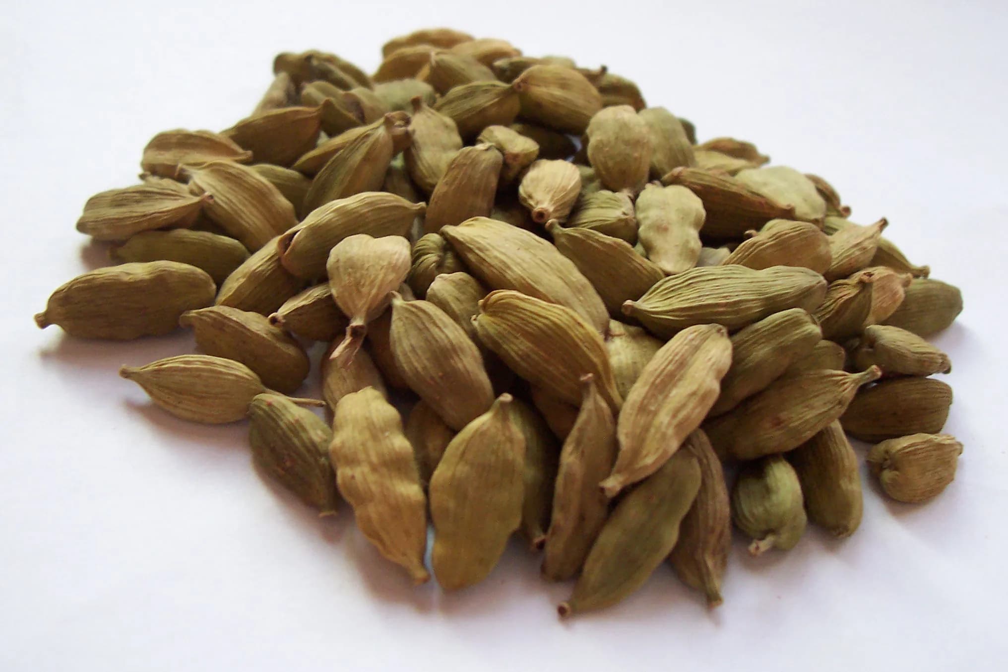 A heap of whole, dried green cardamom pods, olive green and ridged, on a white surface