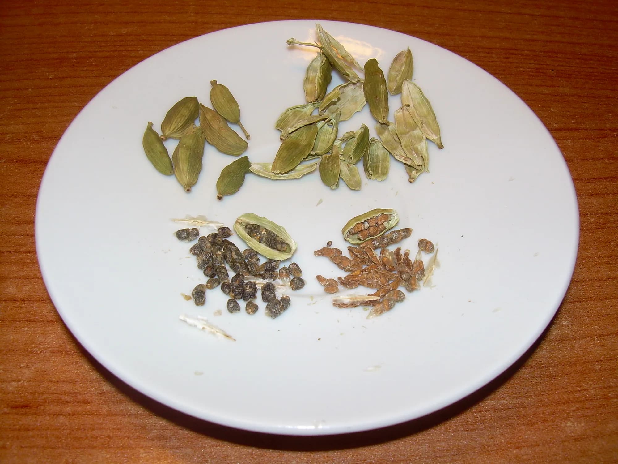 Green cardamom pods on a white plate, whole at the top and split open below with their small dark seeds loose beside them